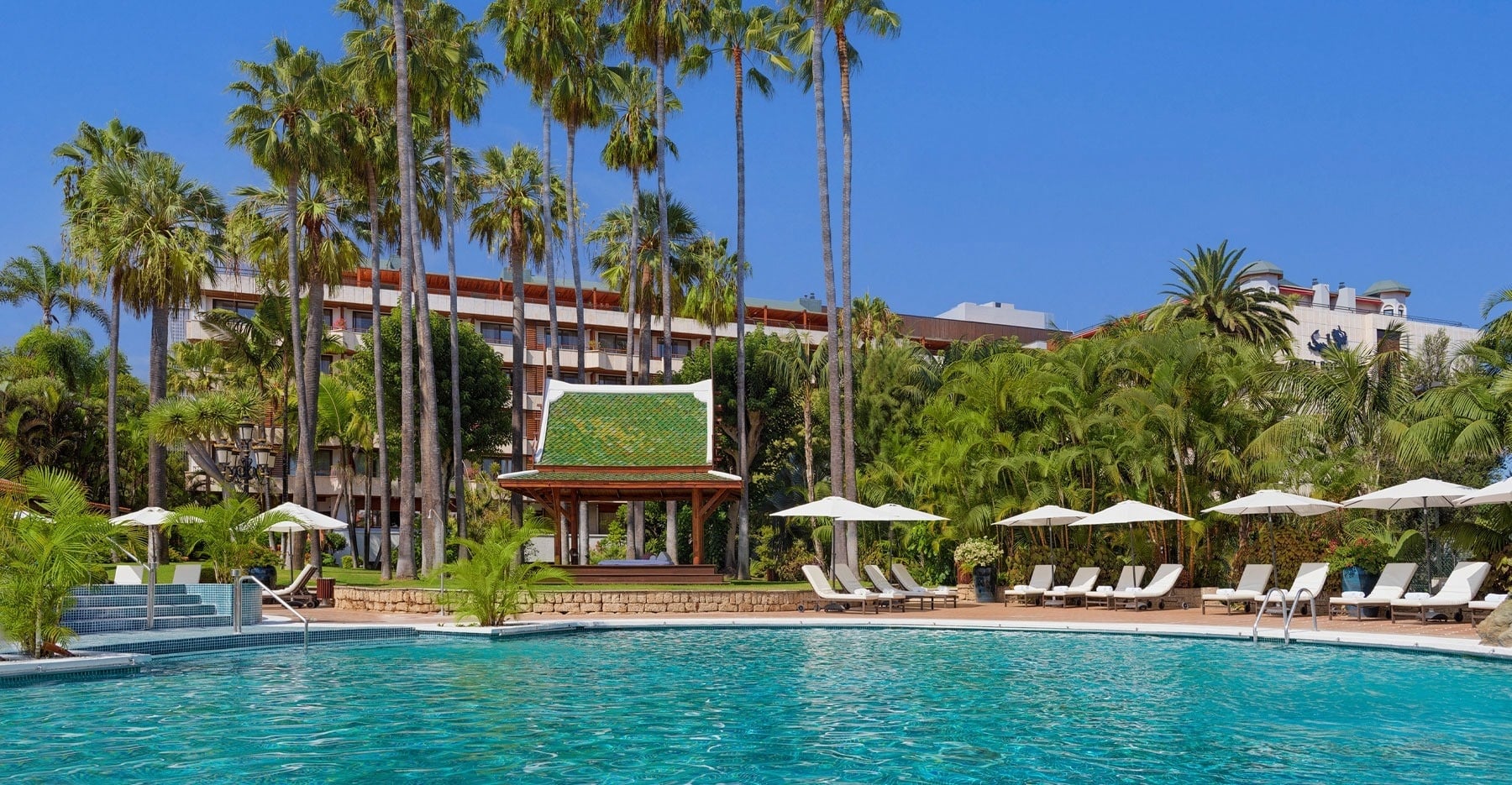 En solig hotellpool omgiven av palmer, en thailändsk paviljong och solstolar, med prislogotyper flytande på vattnet.