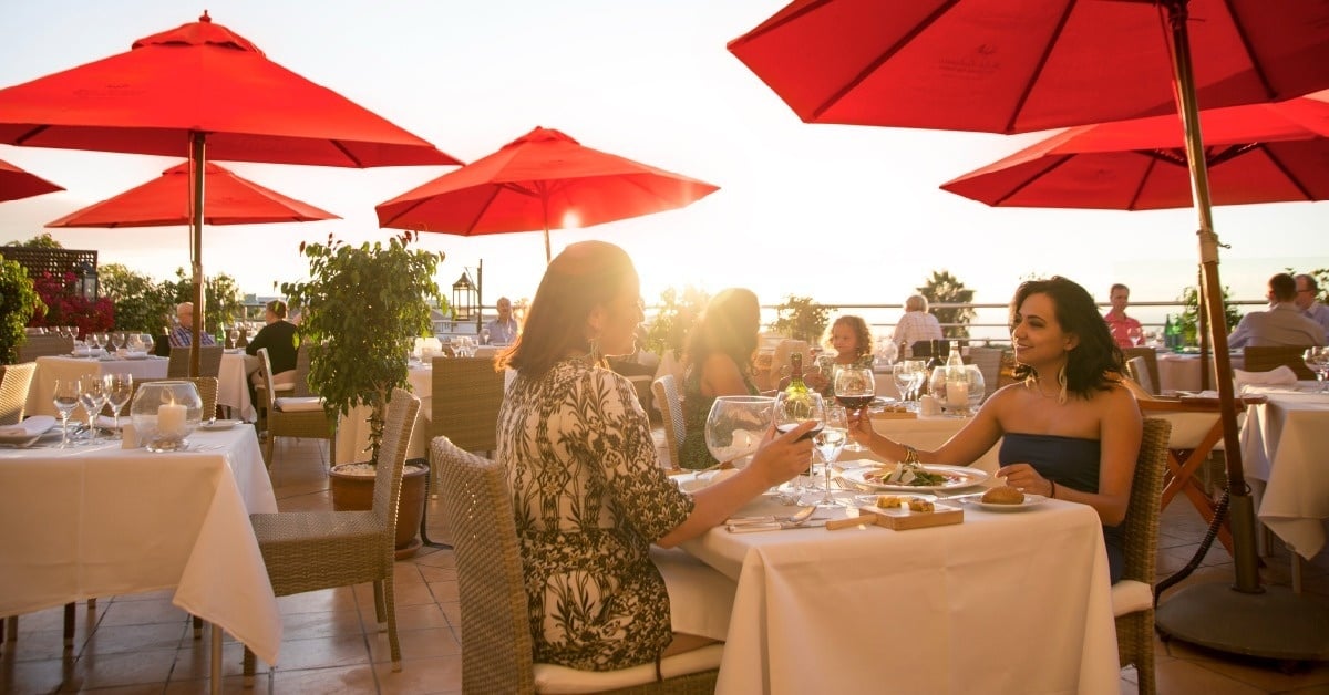 People are enjoying dinner at an outdoor rooftop restaurant with red umbrellas, against the backdrop of a sunset sky.