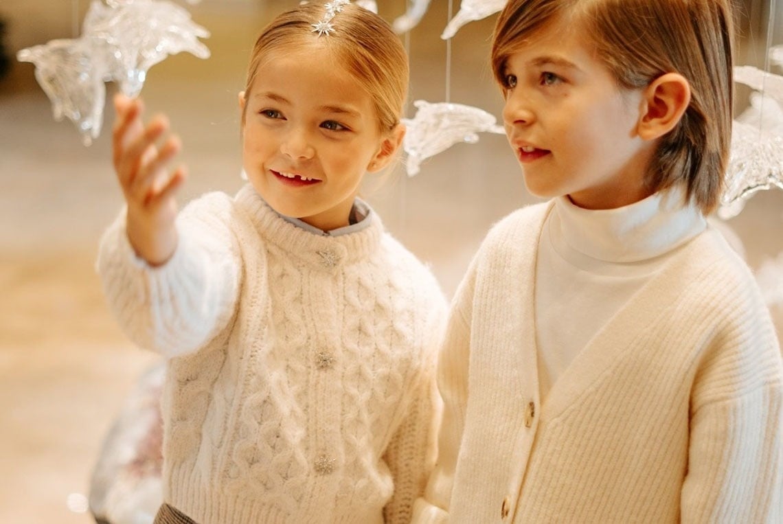 Dos niños, uno sonriendo y el otro mirando, visten suéteres de punto de color crema.