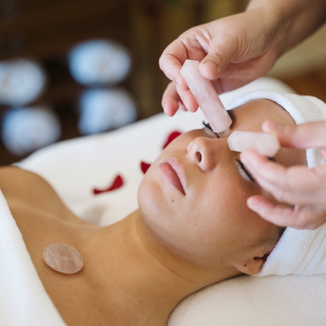 En person får en lugnande spa-behandling med rosenkvartsstenar som placeras på ansiktet och bröstet av en terapeut, omgiven av röda rosenblad.