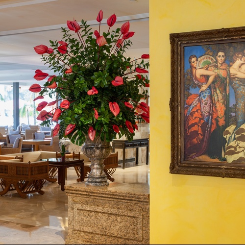 Una grande composizione floreale con anthurium rossi in un vaso di pietra domina il centro di una hall elegantemente arredata, con un quadro incorniciato su una parete gialla sulla destra.