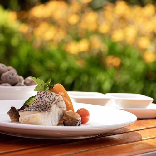 A plate of grilled fish with vegetables and mushrooms is elegantly presented on a wooden table, with a blurred garden background featuring green foliage and yellow flowers.