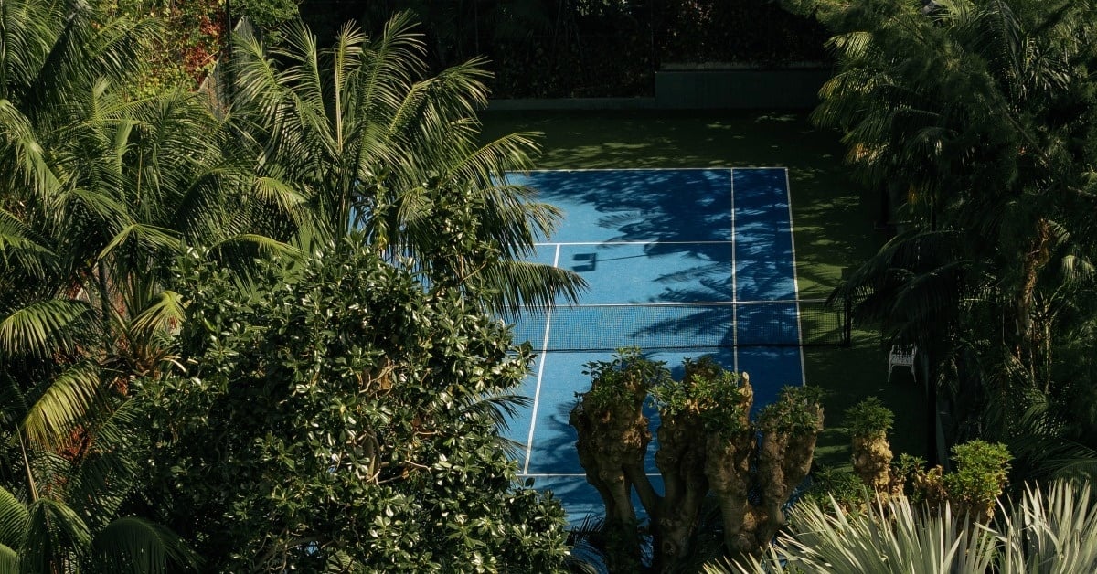 Una cancha de tenis azul rodeada de densa vegetación tropical, incluyendo palmeras, vista desde arriba.