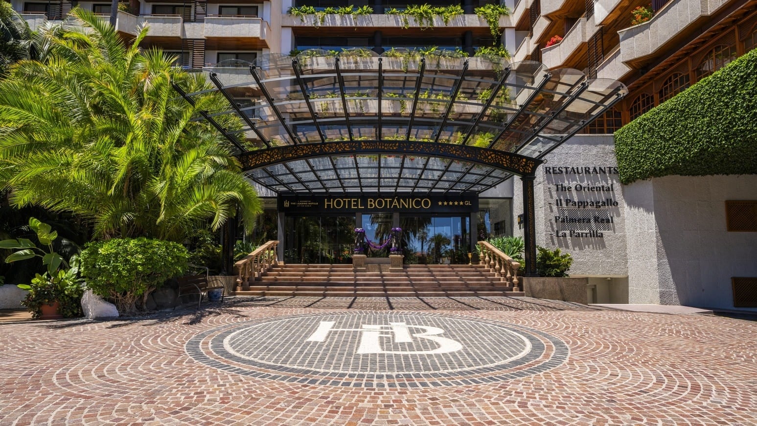 La fachada del Hotel Botánico, rodeada de palmeras y exuberante vegetación, presenta banderas de la UE, Tenerife y España a su entrada, bajo un cielo azul y despejado.
