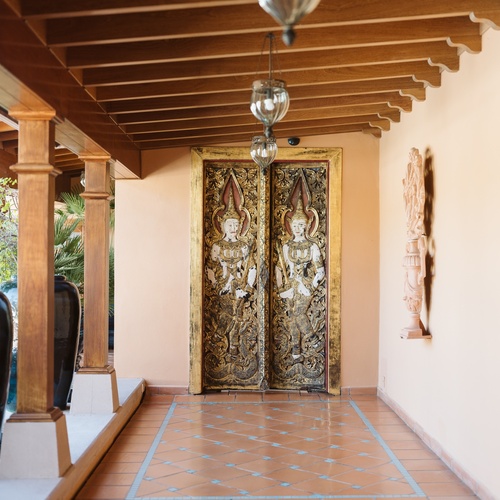 Un corridoio riccamente decorato con un soffitto a travi di legno, porte intagliate con figure asiatiche e un pavimento piastrellato a motivi geometrici, illuminato da lanterne appese.