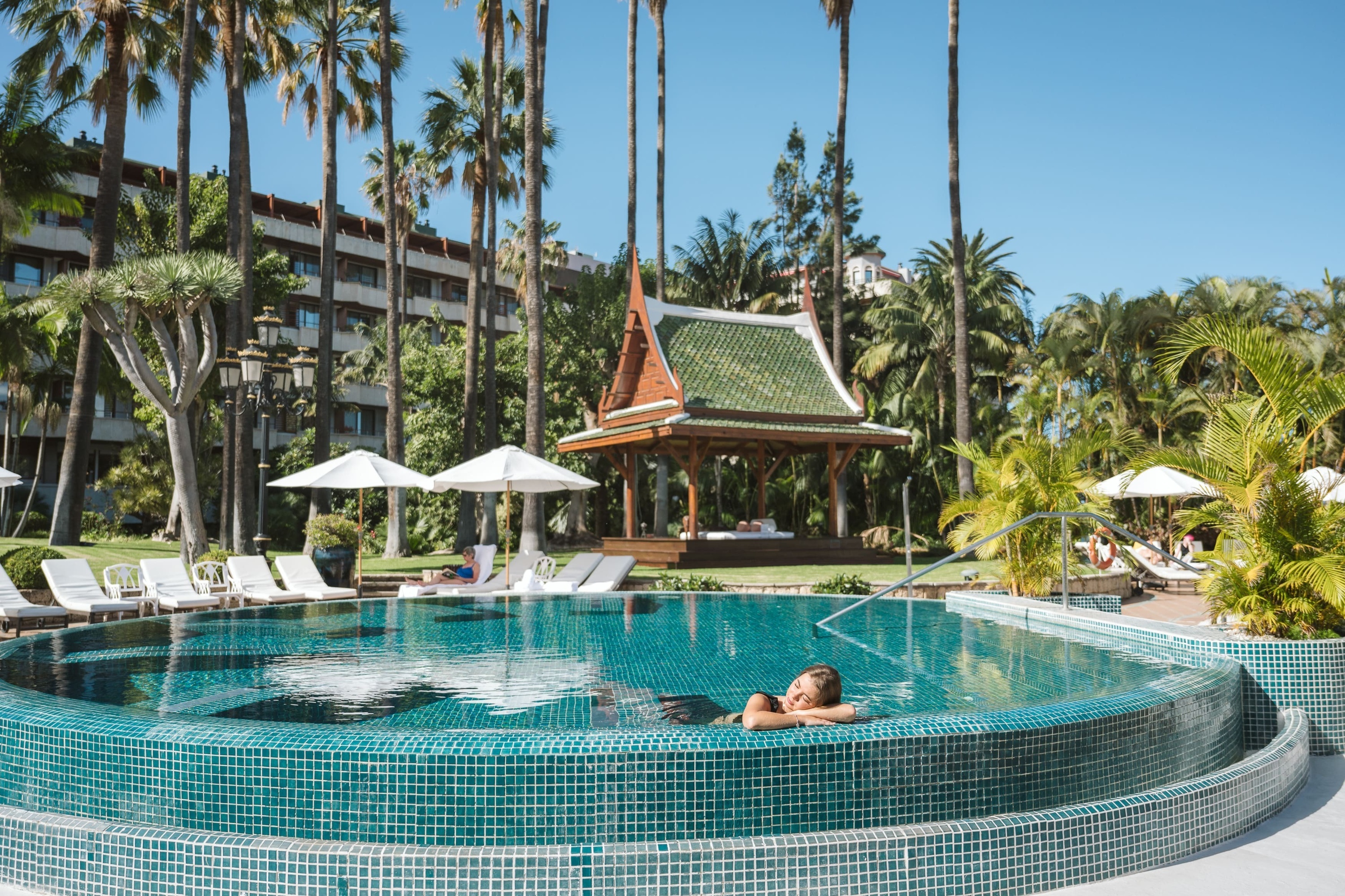 A woman relaxes in a blue-tiled swimming pool at a tropical resort, surrounded by palm trees, a hotel building, and a traditional Thai-style gazebo.