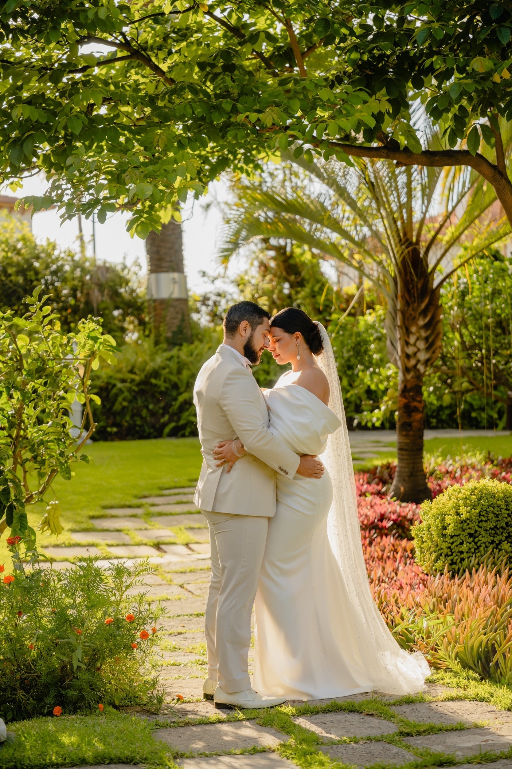 Ett brudpar står och omfamnar varandra ömt under ett lummigt grönt träd i en solbelyst trädgård.
