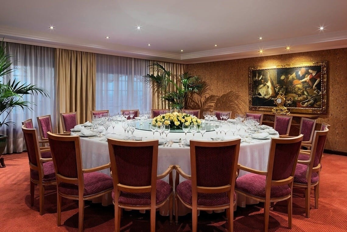 Ett elegant dukat runt middagsbord med många stolar, ett blomsterarrangemang, gardiner, växter och en konsttavla syns i en formell matsal.