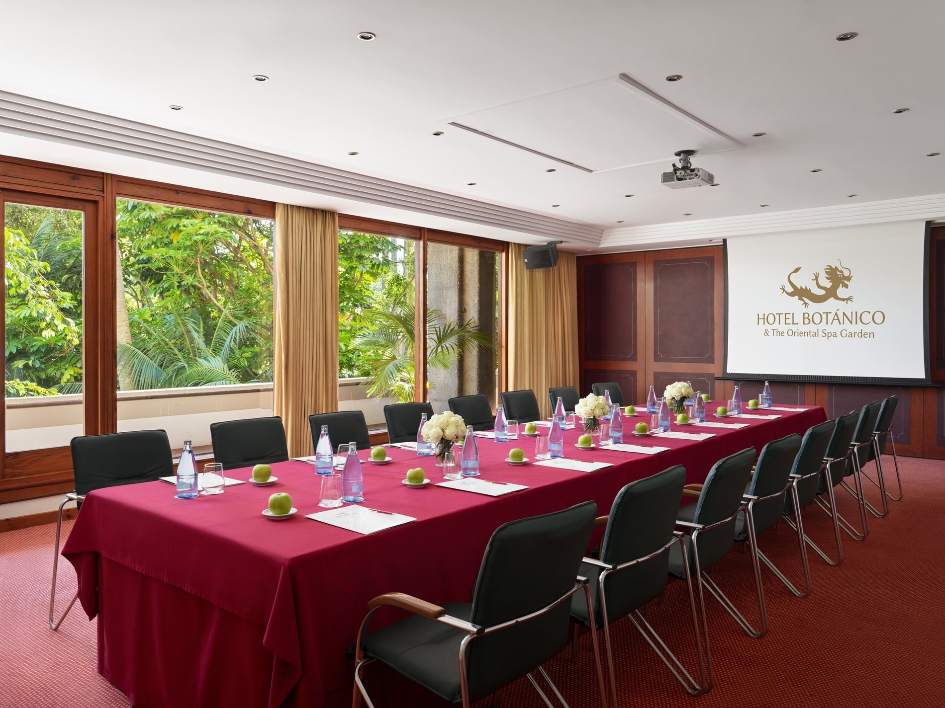 Une salle de réunion élégante est aménagée avec une longue table rouge dressée pour une conférence, faisant face à un écran de projection affichant le logo d'un hôtel et offrant une vue sur un jardin luxuriant à travers de grandes fenêtres.