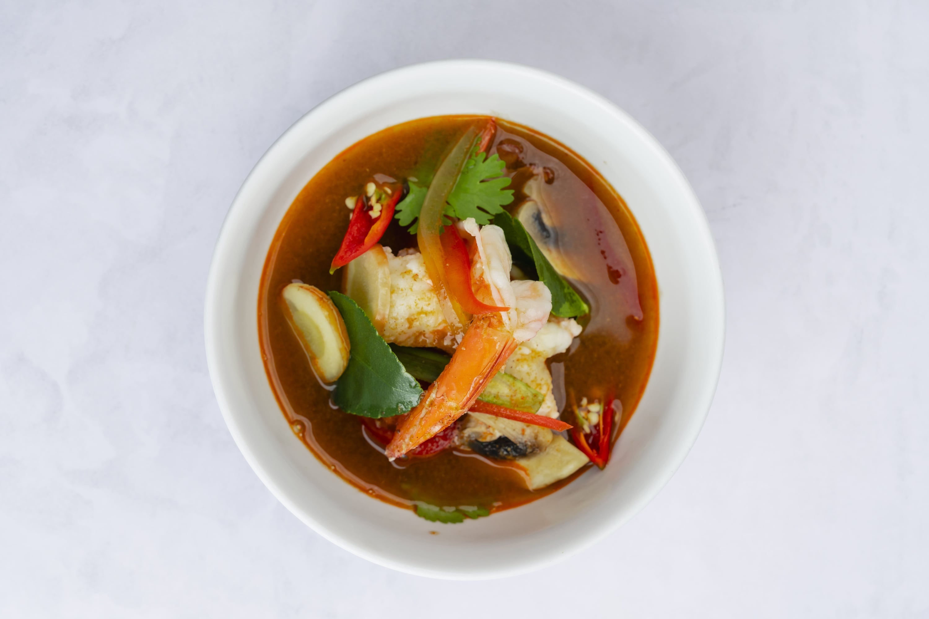 A smiling Asian woman wearing a red chef's jacket, black chef's hat, and glasses stands with her arms crossed, looking at the camera in an indoor setting with a blurred background showing a window and table.
