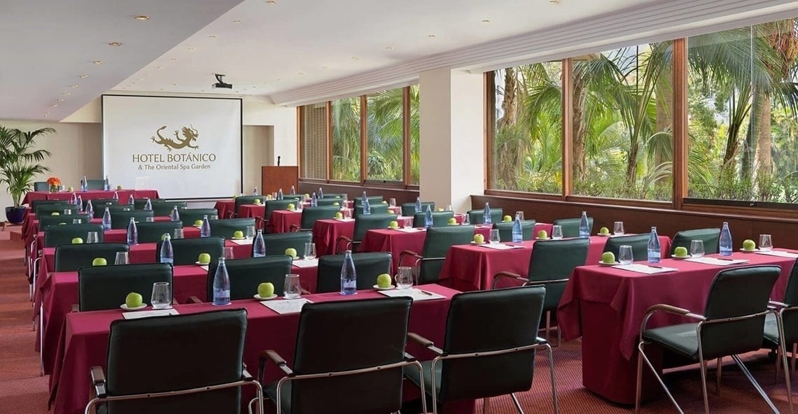 Ett konferensrum är dukat för möte med rader av bord, stolar, vatten och äpplen, med en projektorduk som visar "Hotel Botánico" och stora fönster med utsikt över en grönskande trädgård med palmer.