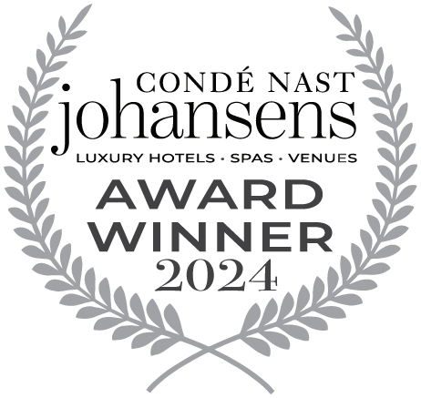 En grå och svart logotyp med en lagerkrans som omger texten "CONDÉ NAST JOHANSENS LUXURY HOTELS • SPAS • VENUES AWARD WINNER 2024".