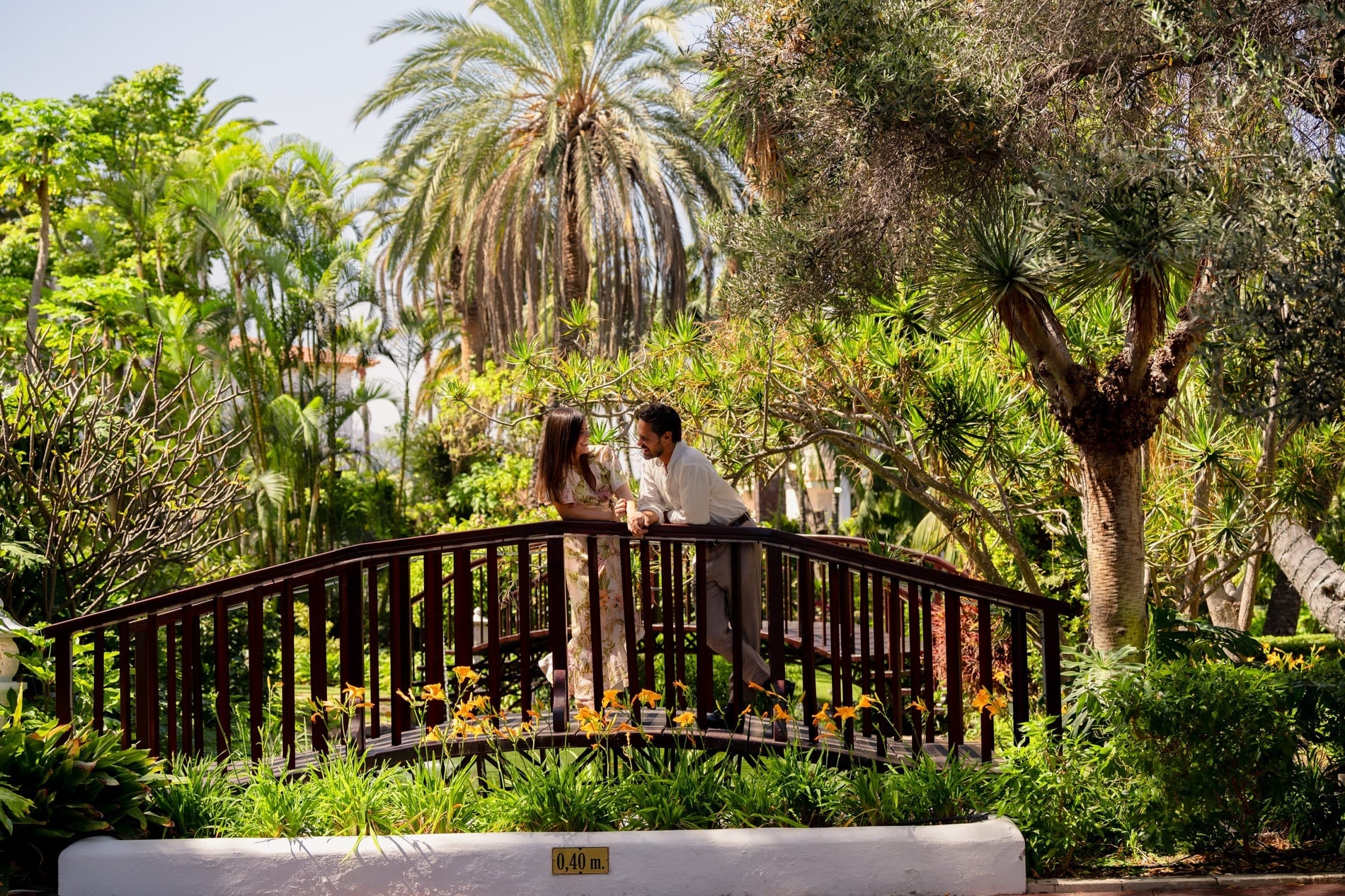 A smiling couple stands on a wooden bridge, looking at each other amidst a vibrant tropical garden filled with palm trees and other lush greenery.
