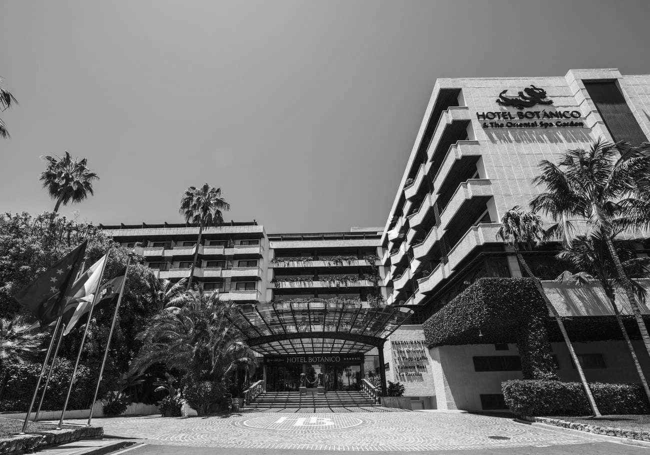 En svartvit bild visar Hotel Botánico, en stor hotellbyggnad omgiven av palmer, frodig grönska och med flaggor vid entrén.=s1900