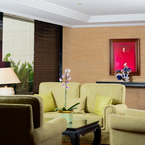 it: Un salotto luminoso è arredato con divani e poltrone di colore verde chiaro, un tavolino di vetro su cui poggia un'orchidea, e presenta un'ampia finestra che rivela la vegetazione esterna, mentre una parete è decorata con carta da parati testurizzata, un quadro rosso e un oggetto decorativo.