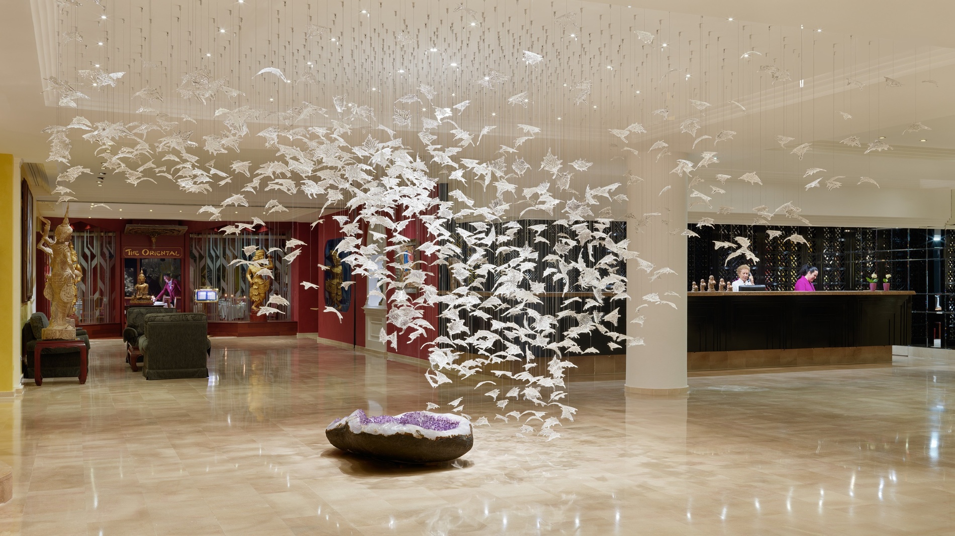 Una elegante hall è dominata da una scultura sospesa composta da centinaia di elementi trasparenti simili a foglie o uccelli, con un grande geode di ametista sul pavimento lucido e una reception sullo sfondo.