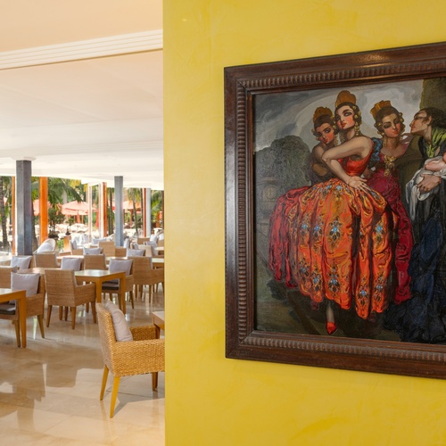 Un dipinto incorniciato con figure colorate è appeso su una parete gialla strutturata, affiancando una luminosa sala ristorante con mobili in vimini e vista su un'area esterna con palme e ombrelloni rossi.