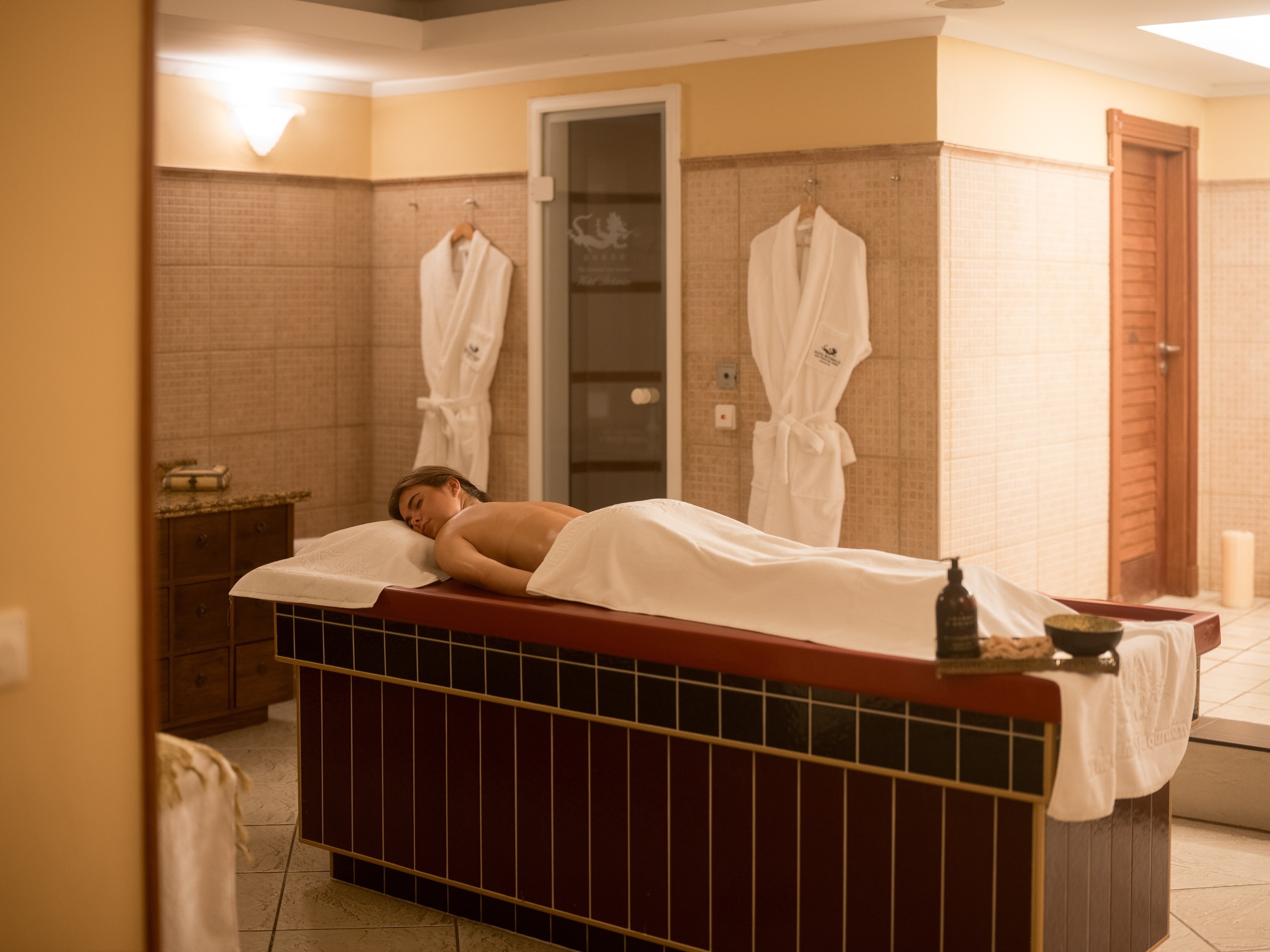 Eine Frau liegt entspannt auf einer Massageliege in einem warm beleuchteten Spa-Raum, umgeben von Bademänteln und Wellness-Accessoires.