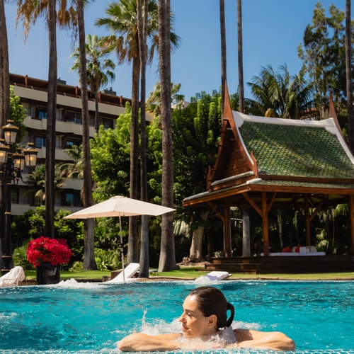 A woman smiles while relaxing in a resort swimming pool surrounded by tall palm trees and a traditional wooden gazebo.