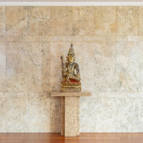 Una statua dorata di Buddha è posta su un piedistallo di pietra contro una parete di marmo chiaro, con un pavimento in legno lucido in primo piano.