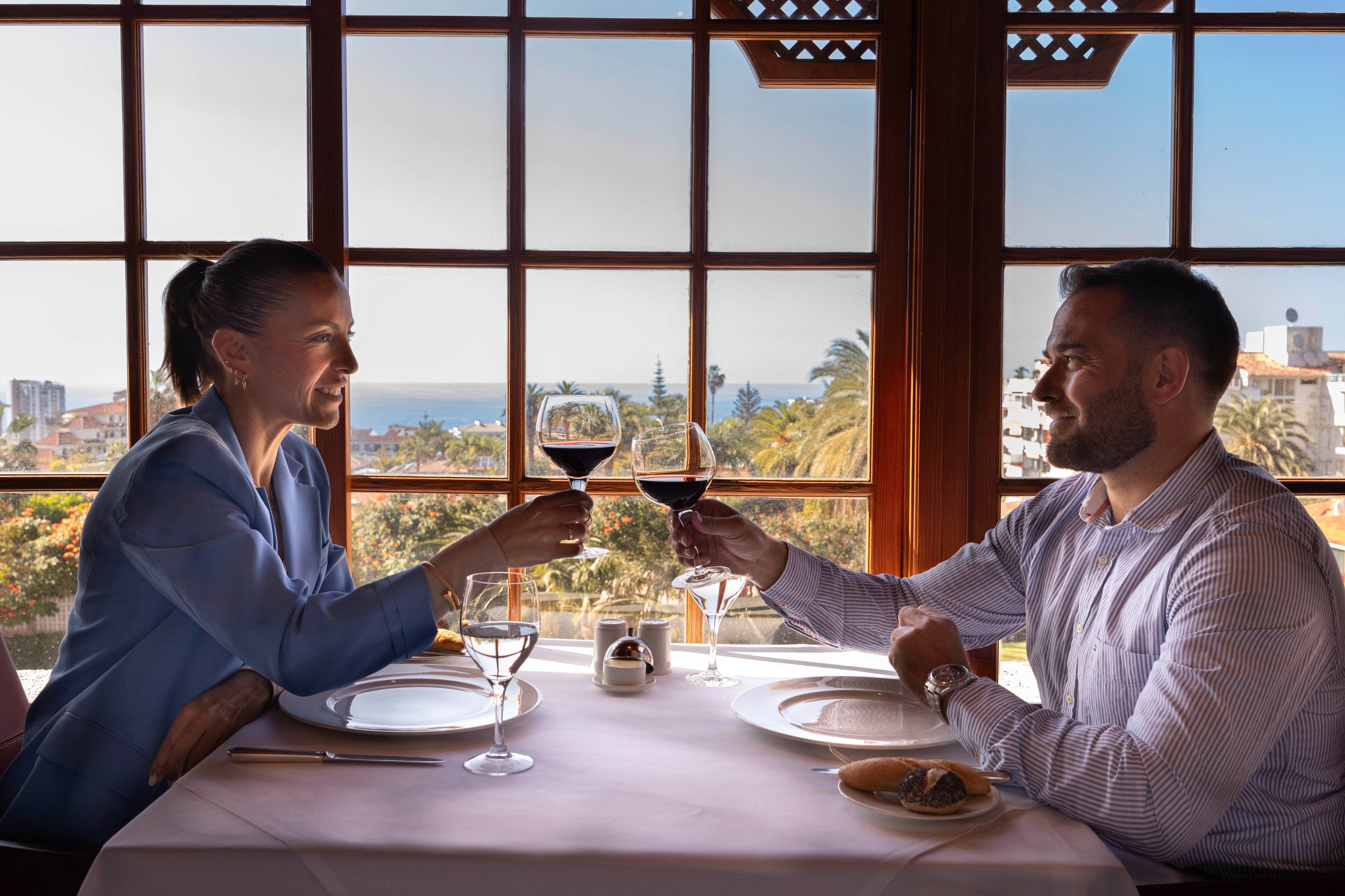 Un hombre barbudo y sonriente sostiene un trozo de postre de chocolate en un tenedor, con una copa de vino tinto en primer plano.