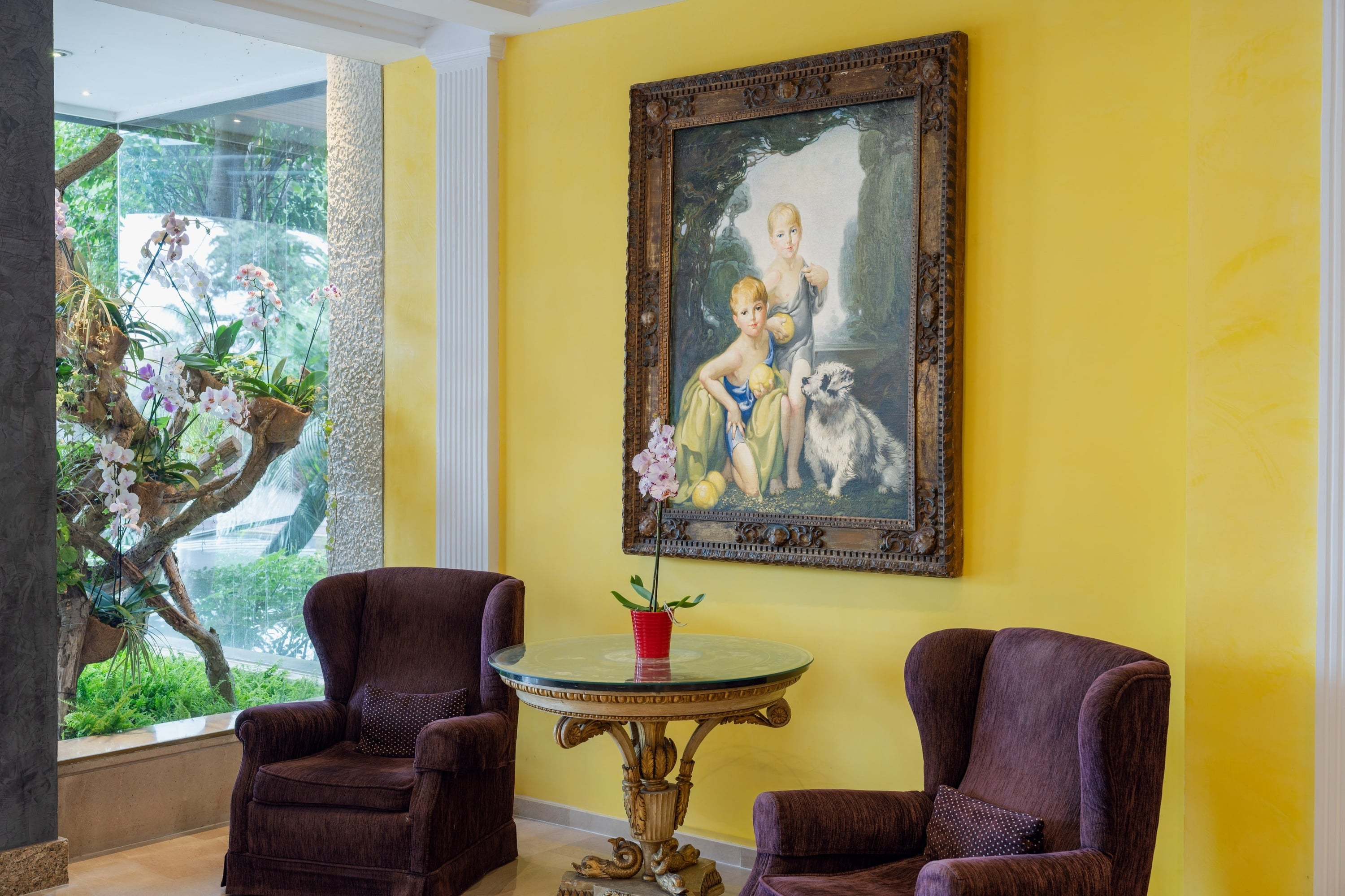 En elegant inredning med ljusgula väggar, ett stort fönster som visar frodig grönska och orkidéer, två mörklila fåtöljer, ett snidat bord med glasplatta och en stor inramad tavla föreställande barn och en hund.