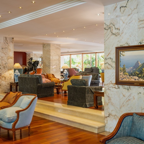 Una elegante lounge di hotel è caratterizzata da pareti in marmo, pavimenti in legno lucido e diverse aree salotto, con un quadro paesaggistico appeso e ampie finestre che mostrano una ricca vegetazione esterna.