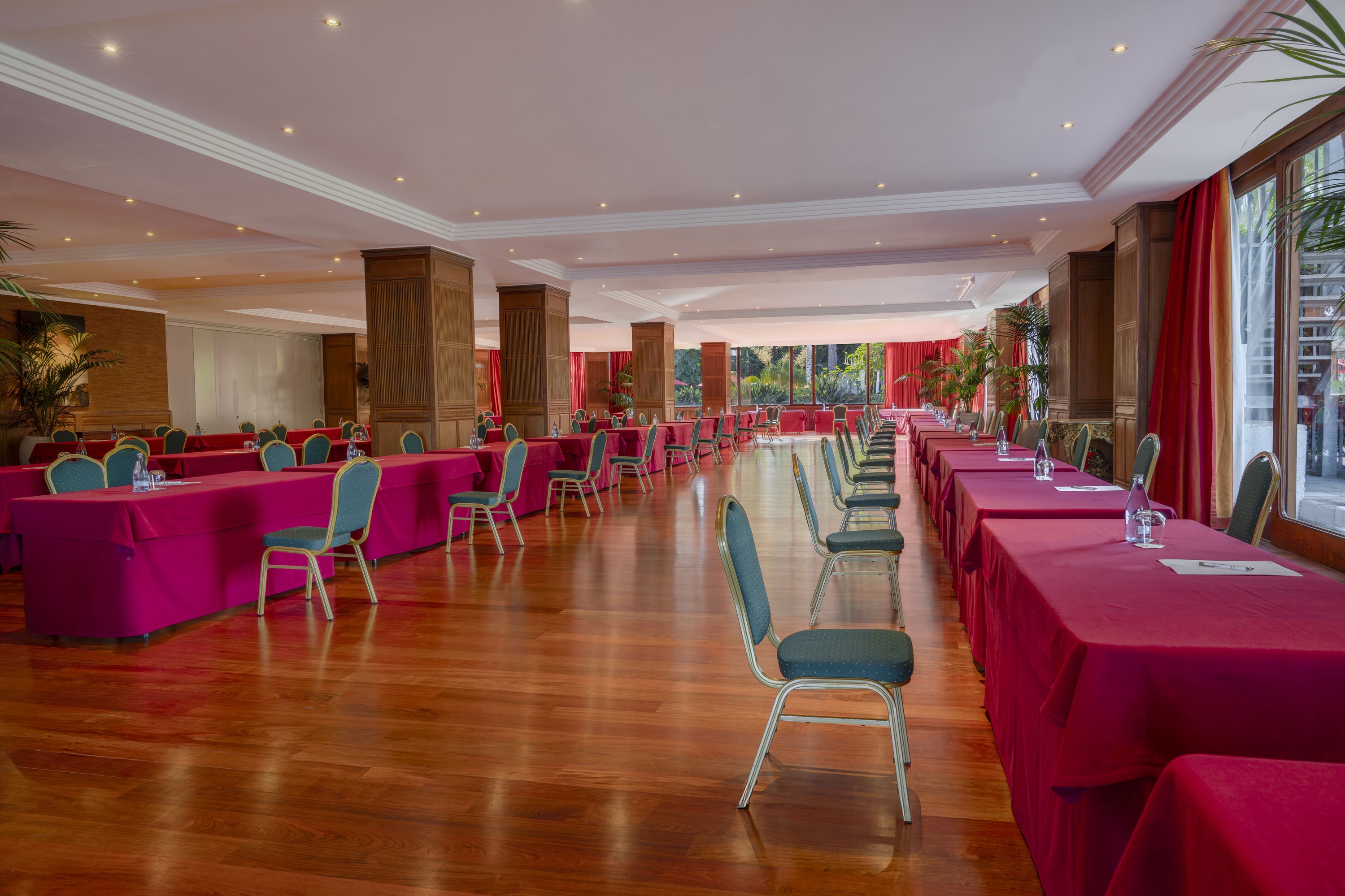 Una espaciosa sala de conferencias está decorada con mesas cubiertas de rojo, sillas con tapicería azul, un suelo de madera brillante y grandes ventanales con cortinas rojas.