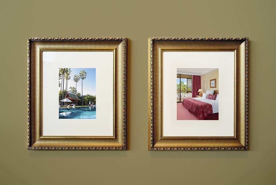Two ornate golden-framed pictures, one displaying a tropical pool area and the other a hotel bedroom, hang side-by-side on a muted green wall.