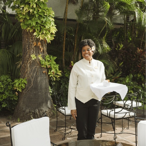 A smiling woman in a cream uniform with a flower in her hair stands outdoors, holding a tray with a white cloth, amidst lush greenery and patio furniture.