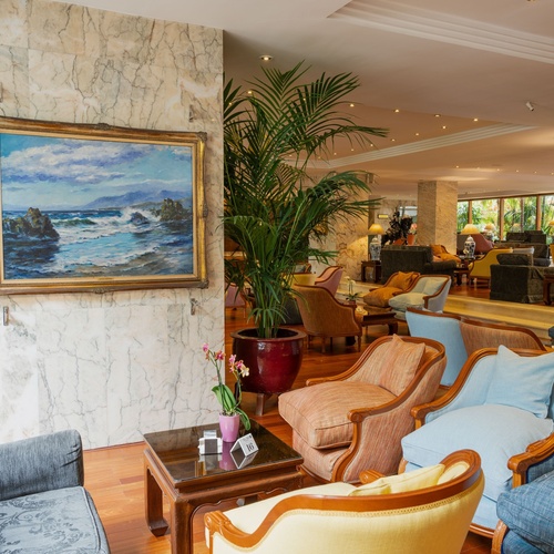 Una lussuosa sala d'attesa o hall d'hotel è arredata con molte poltrone in diversi colori, pavimenti in legno, pareti in marmo e opere d'arte, il tutto illuminato da una luce calda e da piante rigogliose.