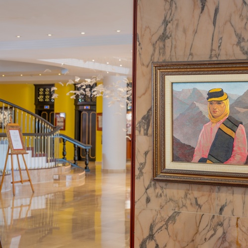 Un dipinto incorniciato raffigurante una figura con abiti tradizionali è appeso su una parete di marmo in una luminosa hall d'albergo con una scala e pilastri sullo sfondo.