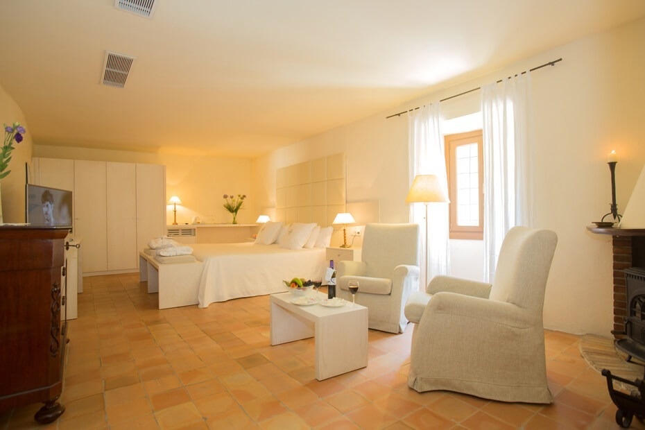 Una amplia y luminosa habitación de hotel presenta una cama doble, una acogedora zona de estar con sillones y una chimenea, todo sobre un suelo de baldosas.