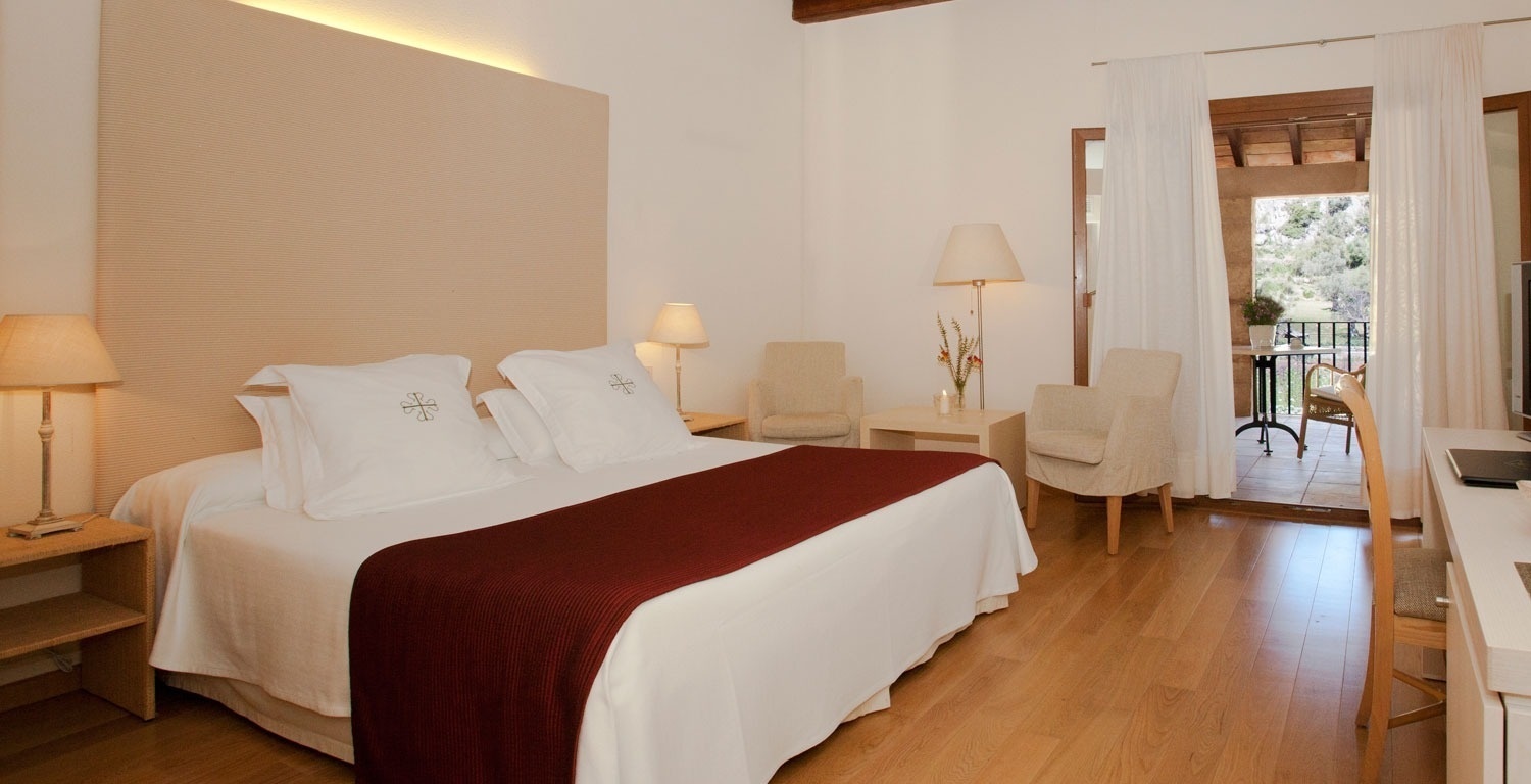 Una luminosa habitación de hotel con cama, sillones, escritorio y un balcón que se abre a un paisaje natural.