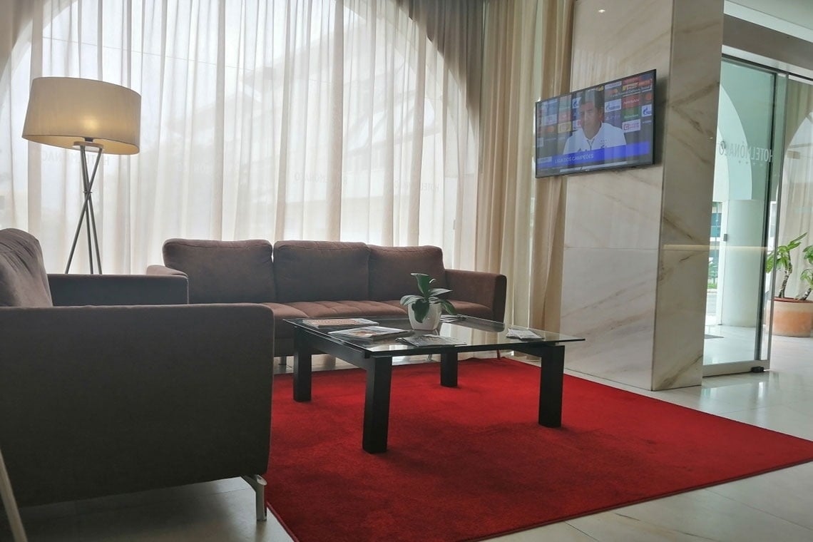 Hotel Mónaco: sala de espera com sofás, mesa de vidro, tapete vermelho e TV.
