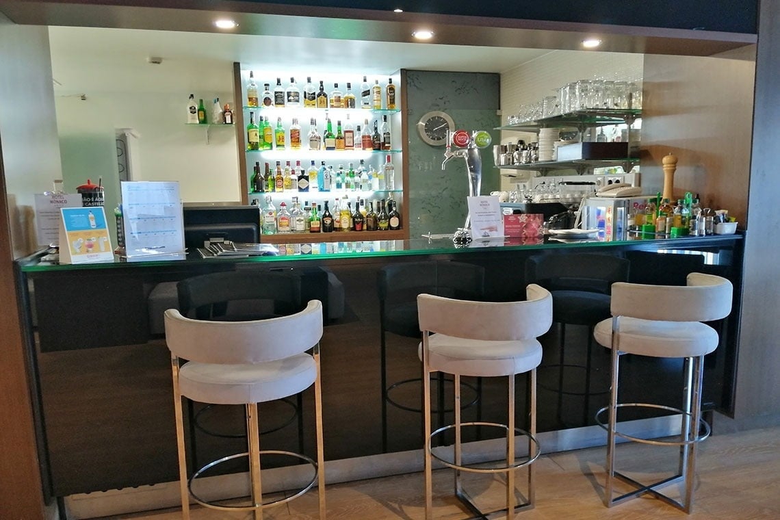 Bar moderno do Hotel Mónaco com bancos altos e garrafas de licor iluminadas.