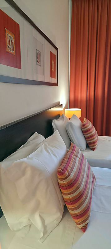 Cama de casal com travesseiros decorativos no Hotel Monaco, com mesas de cabeceira e iluminação moderna.