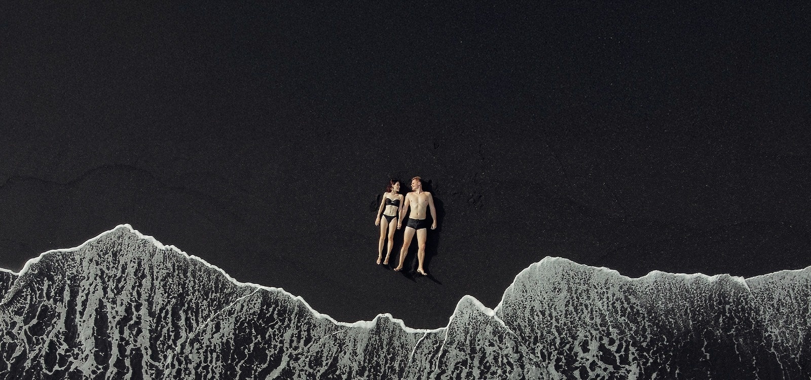 a man and a woman are standing on a black sandy beach