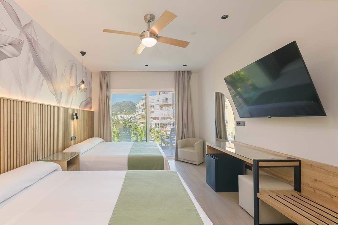 Una moderna habitación de hotel con dos camas, decoración luminosa, balcón con vistas a la ciudad y la montaña, y un televisor en la pared.