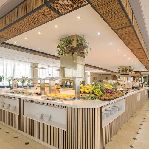 Un amplio y luminoso restaurante tipo buffet presenta una larga barra central con frutas frescas y varios platos, rodeado de áreas de comedor y un techo decorado con elementos de bambú.