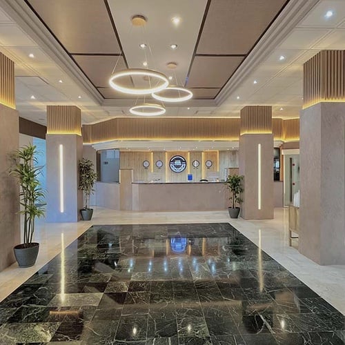 El moderno vestíbulo de un hotel destaca por su mostrador de recepción, pilares con iluminación integrada, lámparas circulares colgantes y un suelo de mármol oscuro y brillante.