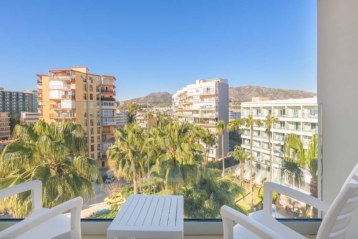 La imagen muestra una vista desde un balcón con sillas y una mesa blancas, mirando hacia una ciudad con palmeras, edificios y montañas al fondo bajo un cielo azul claro.