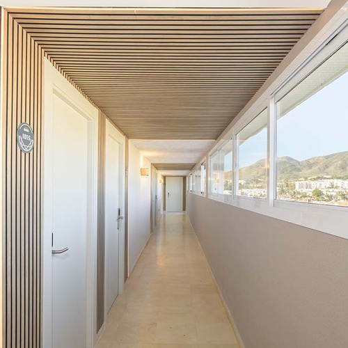 Un largo pasillo de hotel con puertas blancas, un techo de listones de madera y ventanas que ofrecen vistas a un paisaje montañoso con edificaciones.