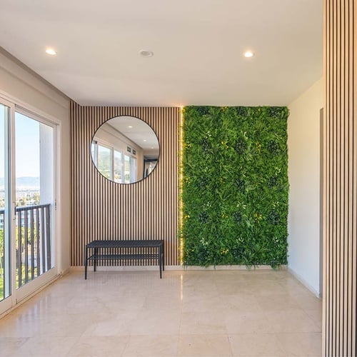 Un pasillo moderno presenta un jardín vertical iluminado junto a una pared de listones de madera con un espejo redondo y un banco, frente a grandes ventanales que ofrecen vistas a la ciudad.