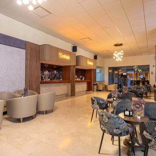 La imagen muestra un amplio y moderno bar de cócteles y zona de lounge con dos mostradores, diversos asientos y suelo de baldosas.