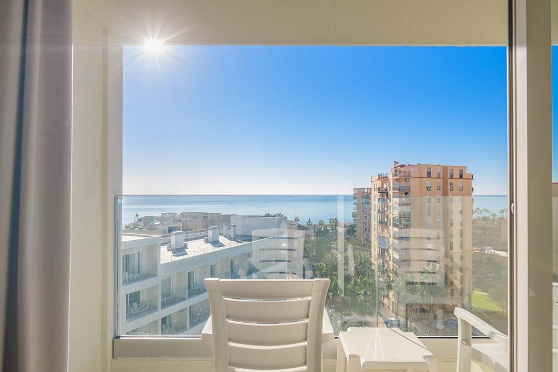 Vista soleada desde un balcón con una silla blanca, que da al mar azul y a edificios bajo un cielo despejado.