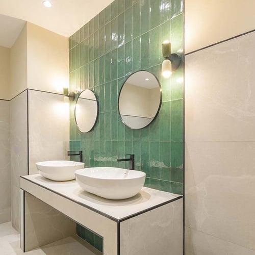 Este es un baño moderno con dos lavabos blancos sobre un tocador de tonos claros y una pared de azulejos verdes brillantes, complementado con espejos redondos y apliques de pared.
