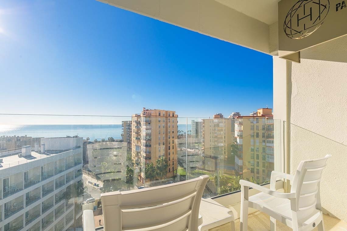 Una vista soleada desde un balcón con el mar azul, edificios de la ciudad y sillas blancas en primer plano bajo un cielo despejado.
