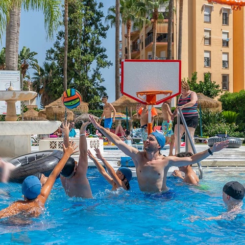 Varias personas juegan animadamente en una piscina con una pelota de voleibol y un aro de baloncesto, rodeadas de palmeras y edificios.