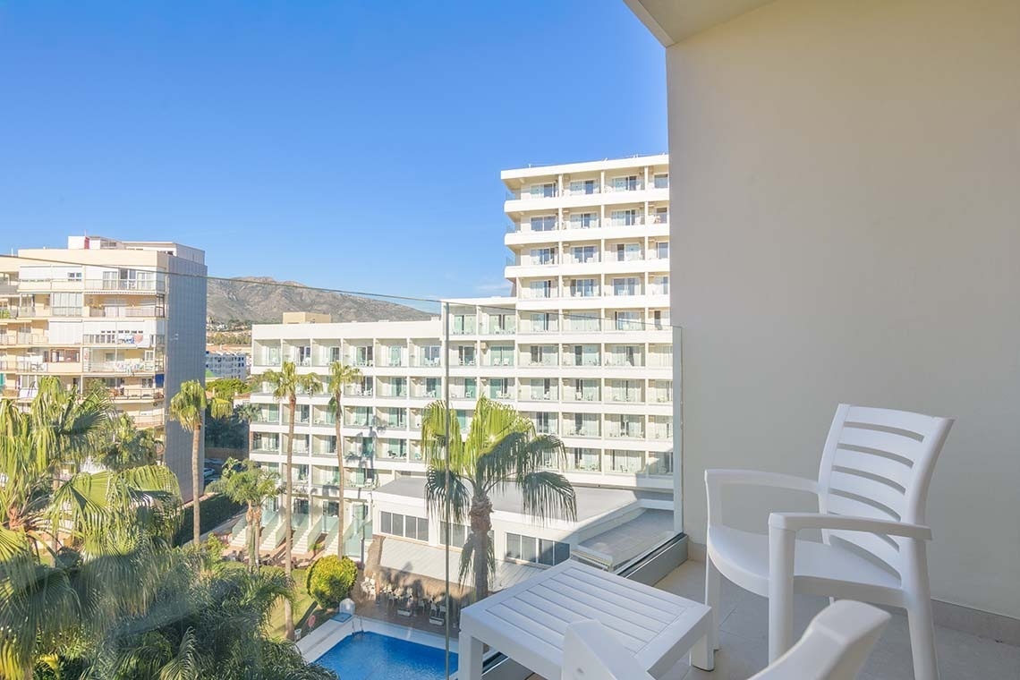 Una vista soleada desde un balcón con muebles blancos, que muestra un complejo hotelero con piscina y palmeras, y montañas al fondo bajo un cielo azul.